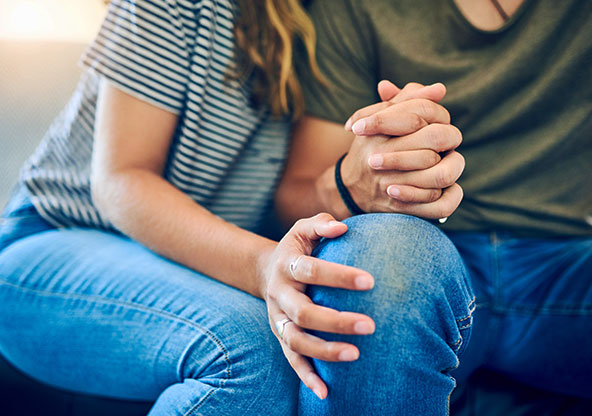 A couple struggling with infertility are holding hands while sitting on the couch at home. 