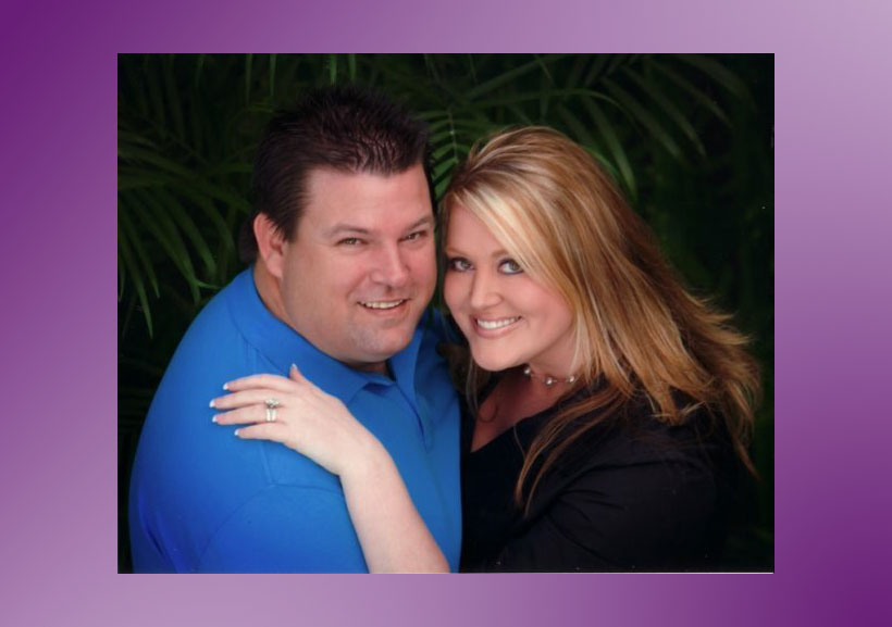 Man in blue shirt with brown hair and woman in black shirt with blonde/brown hair. Both are hugging each other.